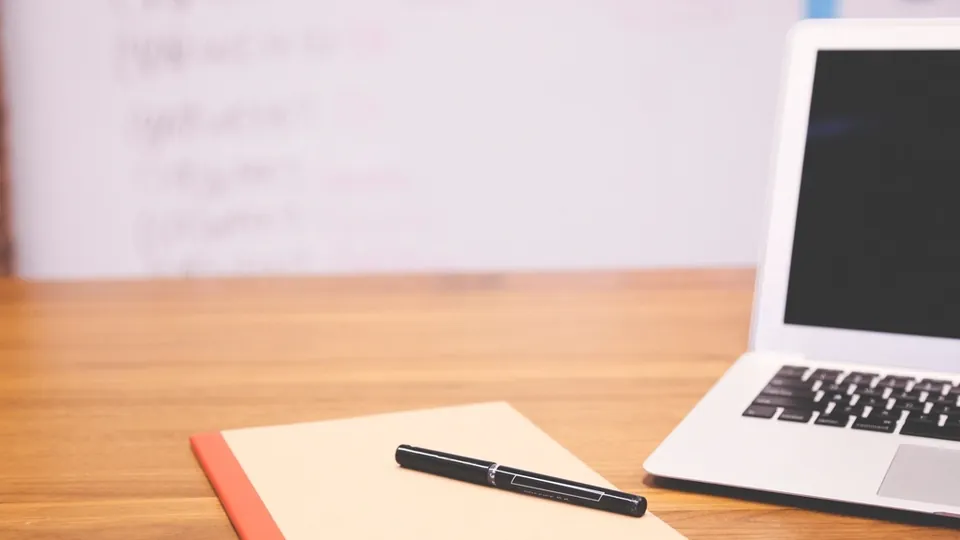 Open notebook and pen on a wooden desk beside a laptop